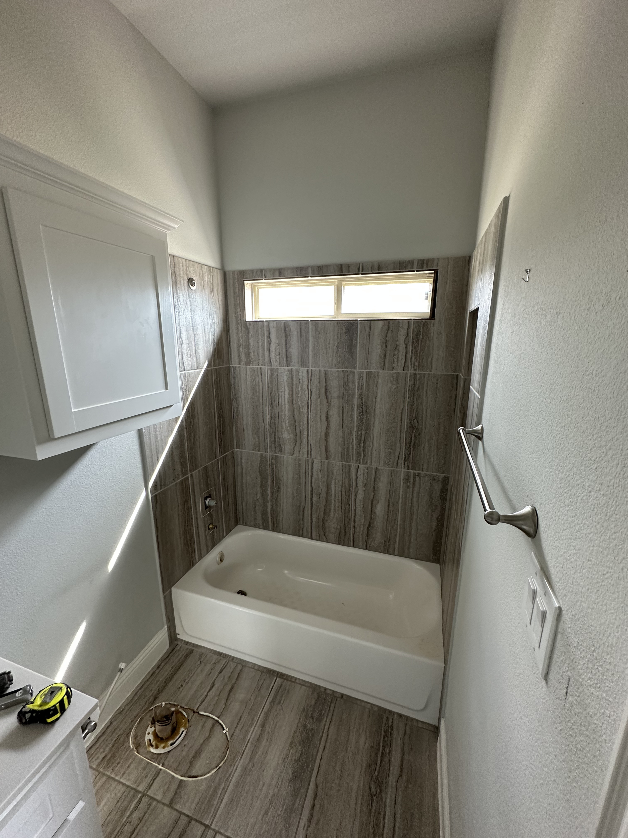 A bathroom under renovation with tiles on the walls and floor, a bathtub, a wall-mounted cabinet, a handrail, and tools on the floor.