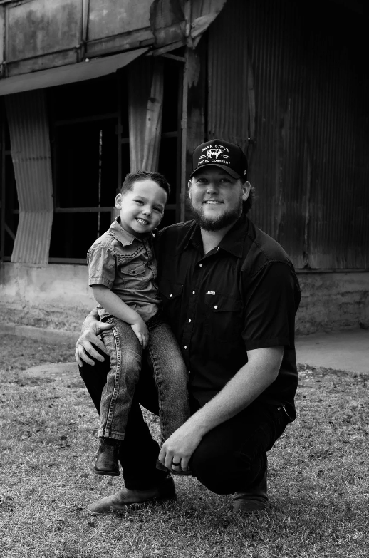 A young boy sitting on an adult man's knee outdoors, both smiling, with a rustic barn in the background.