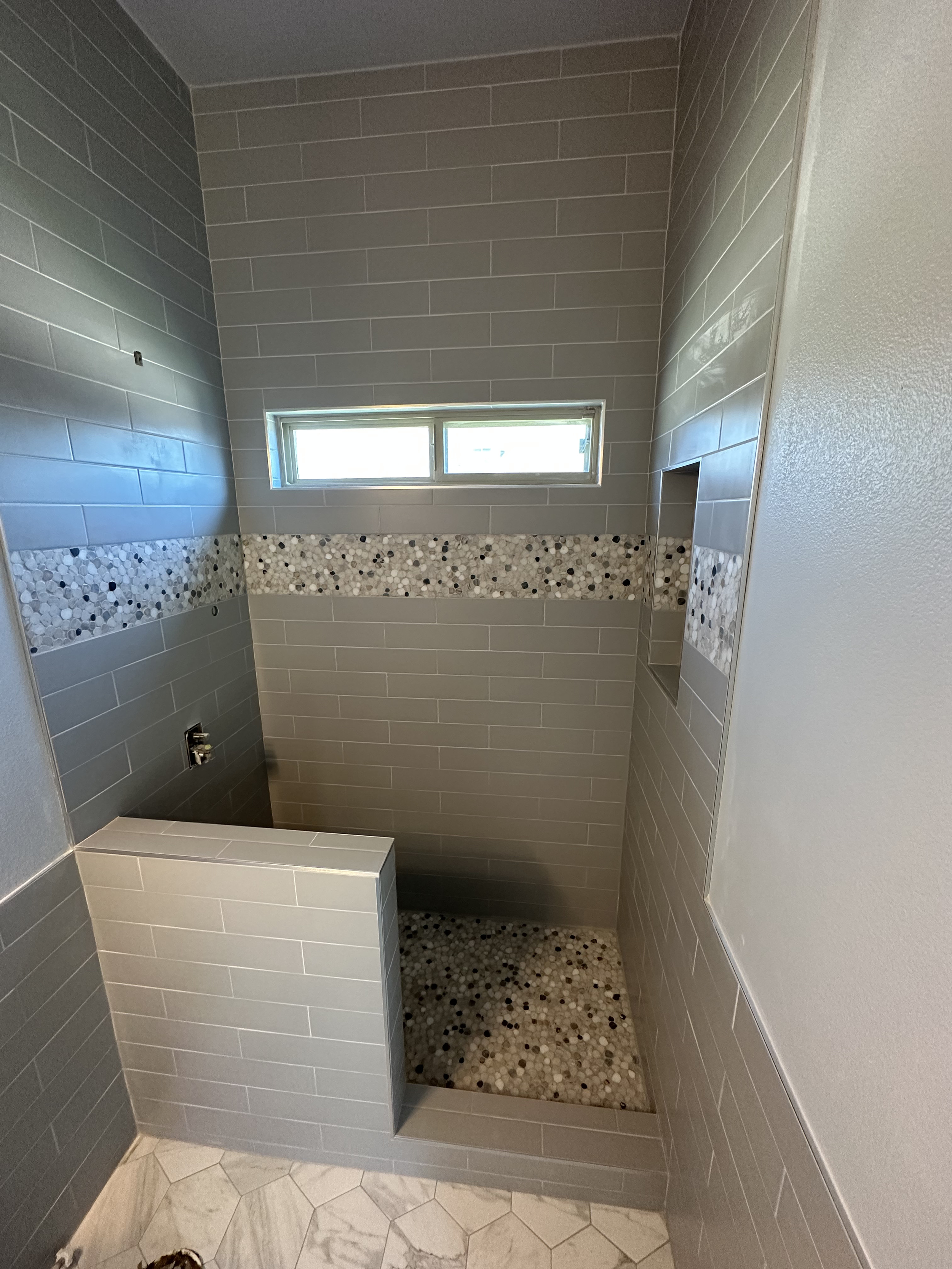 A small bathroom shower area with gray subway tiles, a horizontal strip of pebble mosaic tiles, a rectangular window, and beige hexagonal floor tiles.