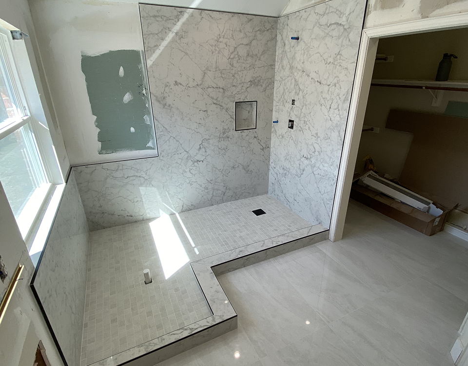 A bathroom shower area under construction with marble tiles on the walls and small square tiles on the floor, a window allowing sunlight in, and an open doorway leading to a walk-in closet.
