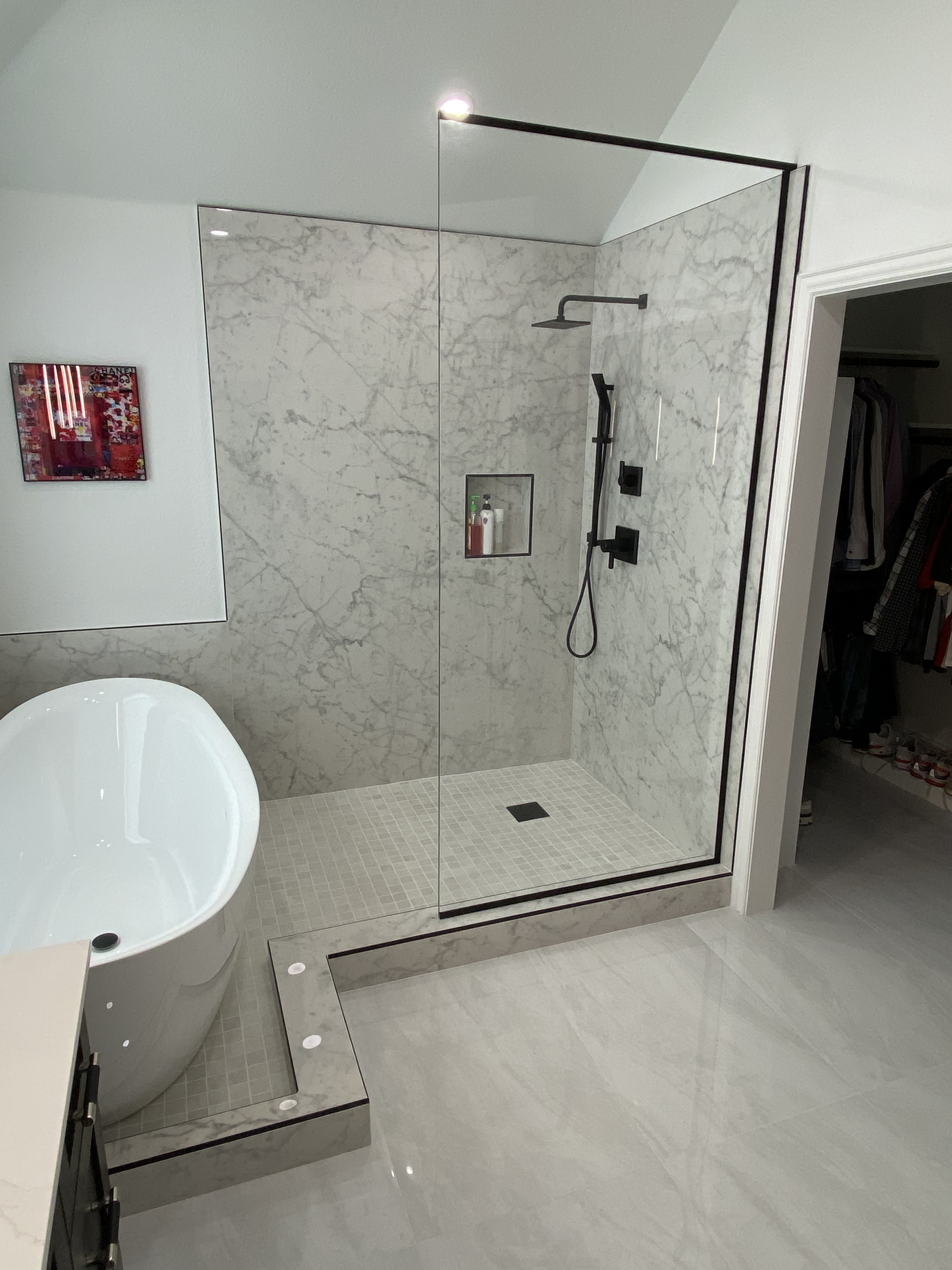Modern bathroom with marble walk-in shower and white oval bathtub. Bathroom wall features a niche with toiletries. Closet with open door visible beside the shower.