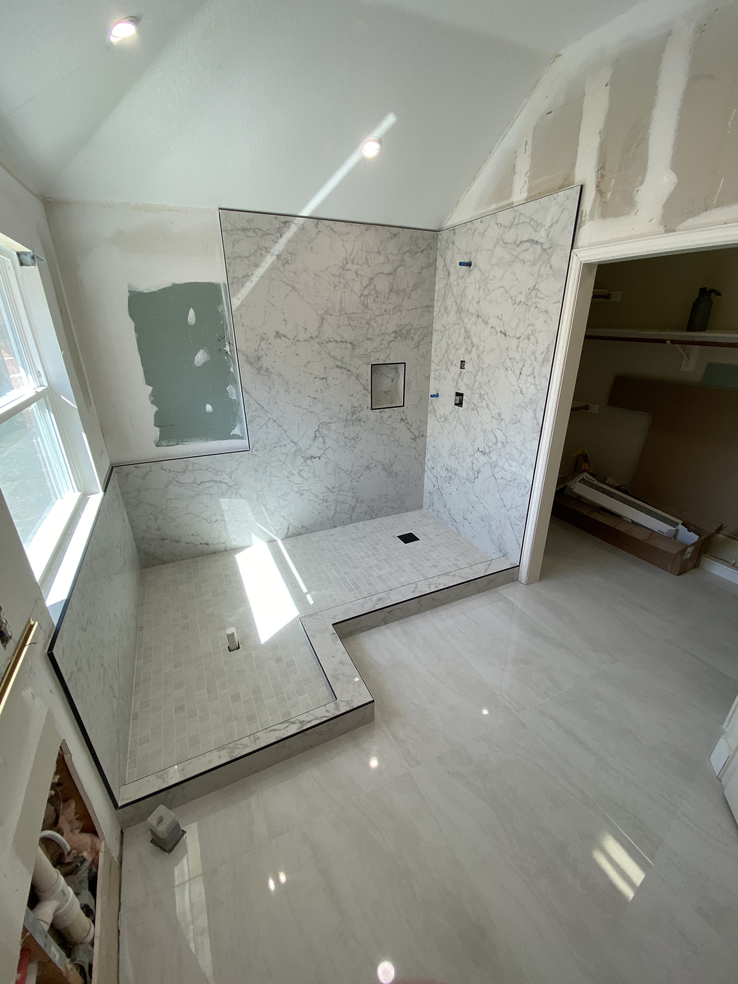 Bathroom under construction with marble walls, a shower area with a small tiled floor, and an empty open closet space on the right. There are construction materials and tools visible.