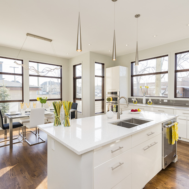 Modern white kitchen with large island, stainless steel sink, and pendant lights, adjacent to a dining area with large windows and natural light.