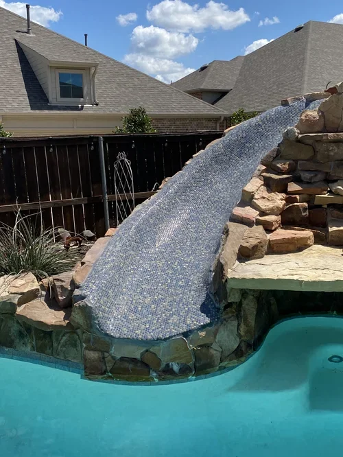 A swimming pool with a small water slide made of rocks and a fabric cover, located in a backyard enclosed by a wooden fence, with neighboring houses and a partly cloudy sky in the background.