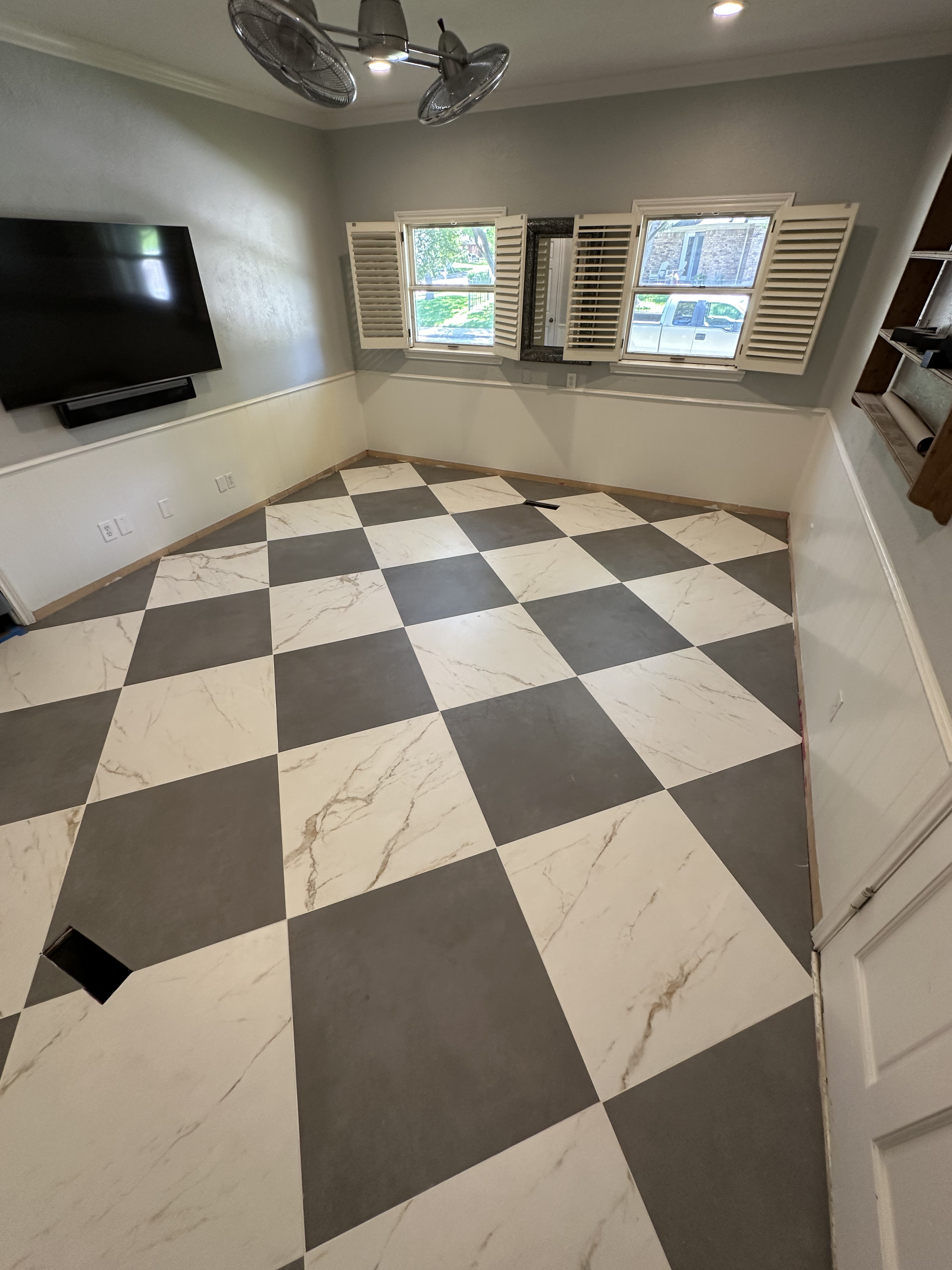 An empty room with a tiled black and white checkered floor, light gray walls, a window with white shutters, and a wall-mounted television.