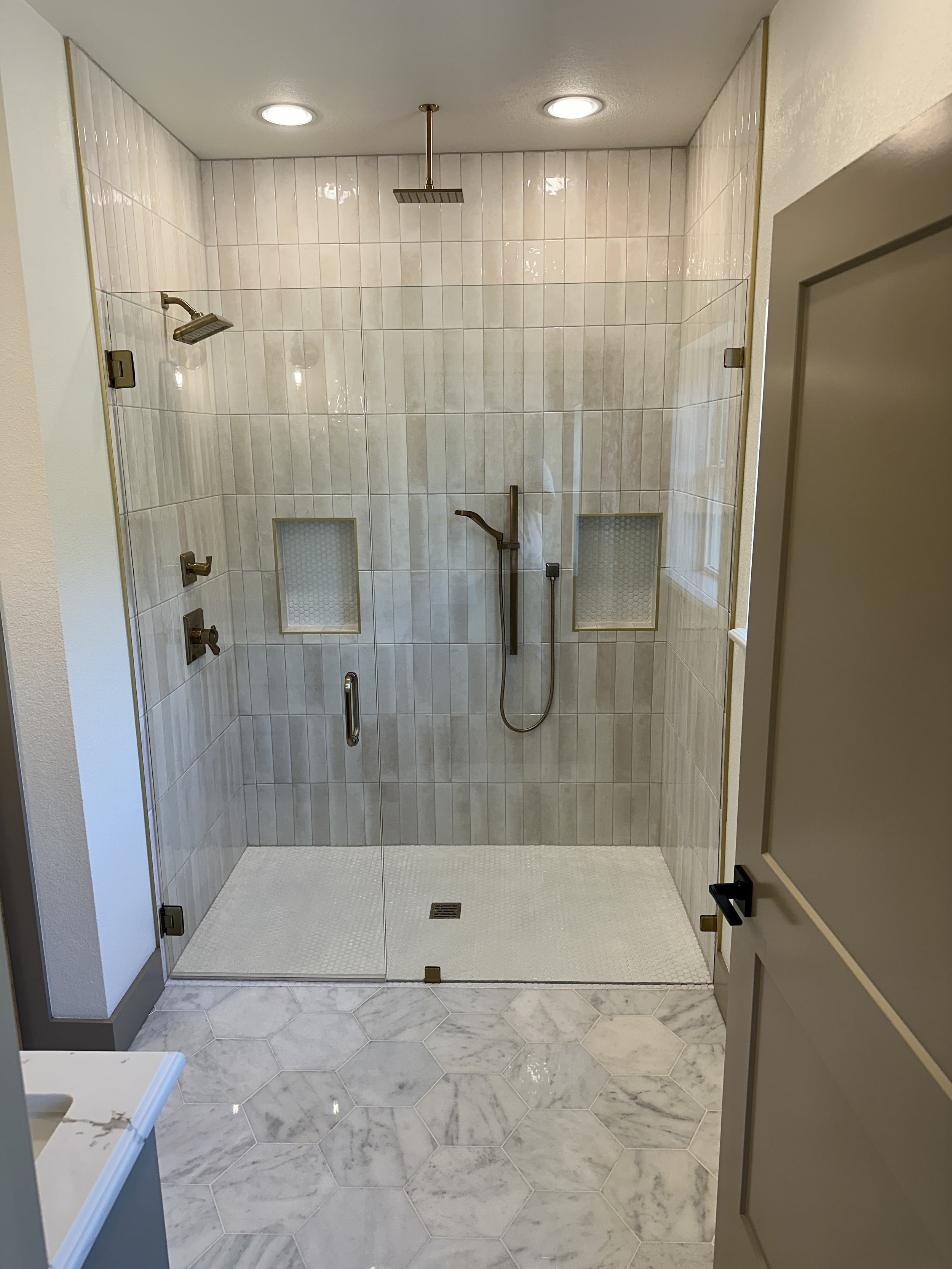 A modern walk-in shower with beige tiled walls, a large ceiling rainfall showerhead, a handheld showerhead, and two recessed tiled shelves, with white hexagonal tile flooring and a glass door.