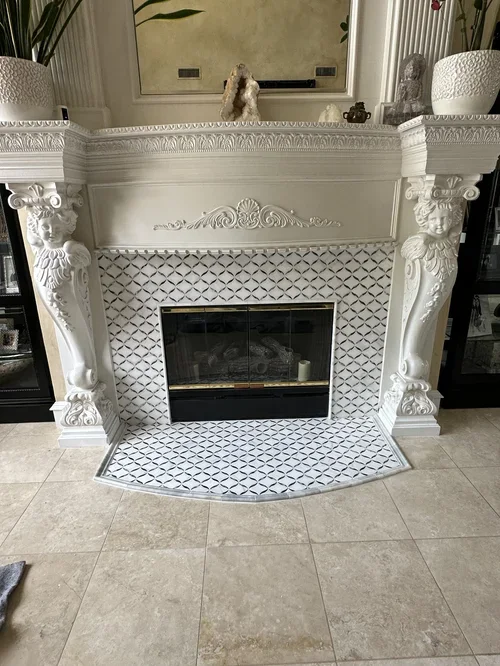 A decorative white fireplace with ornate carved details and columns, surrounded by patterned tiles and flanked by potted plants and decorative items on top.