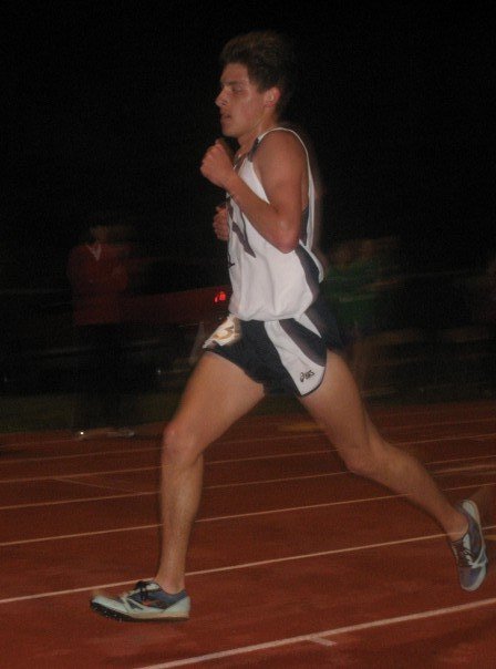 Male athlete running on track at night, wearing athletic shorts, a tank top, and running shoes.