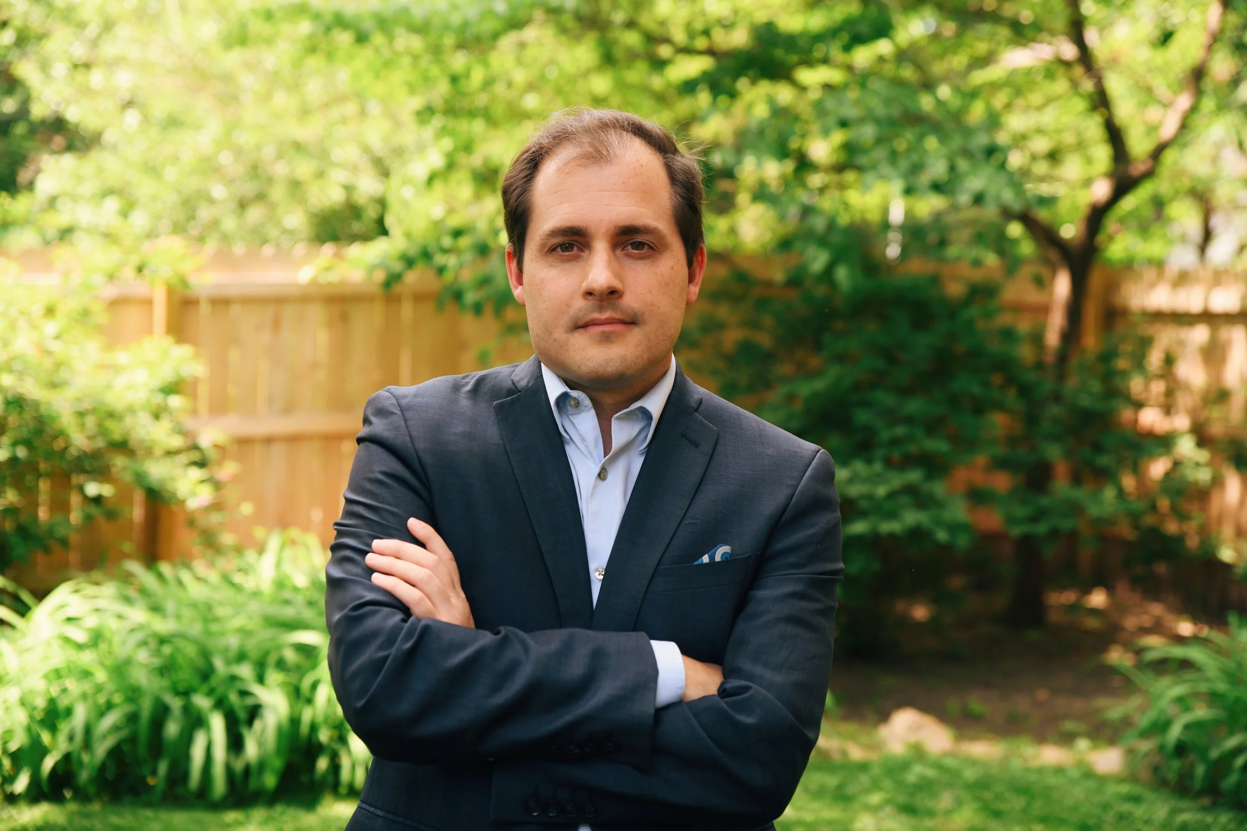 A man in a suit standing outdoors with arms crossed, surrounded by green trees and a wooden fence in the background.