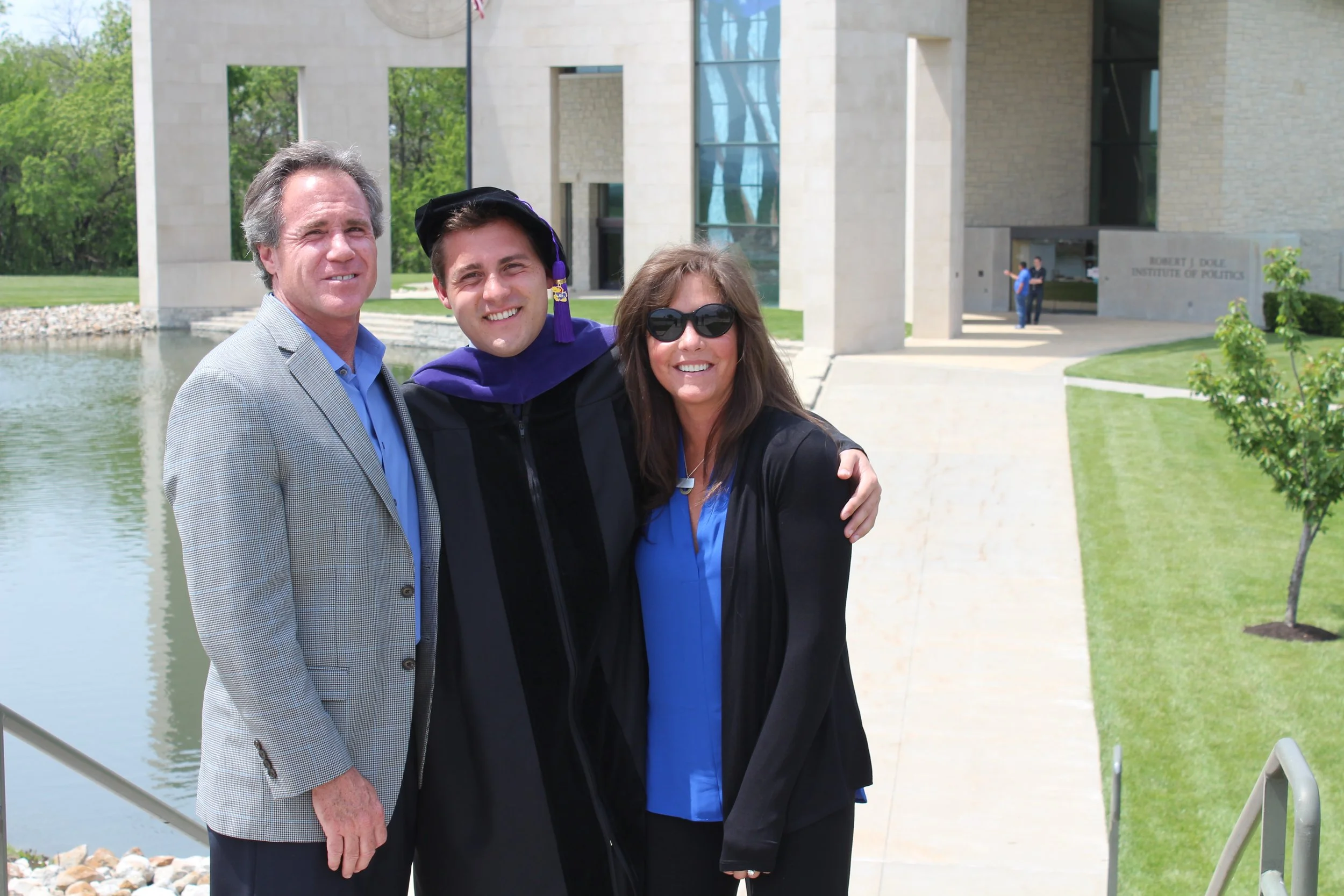 Three people standing outside in front of a modern building with water and greenery surrounding it, celebrating a graduation. The person in the middle is wearing a graduation cap and gown, while the two others are dressed in business casual attire.
