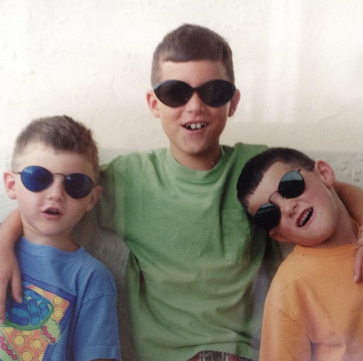 Three boys wearing sunglasses sitting together on a couch, smiling and making fun poses.