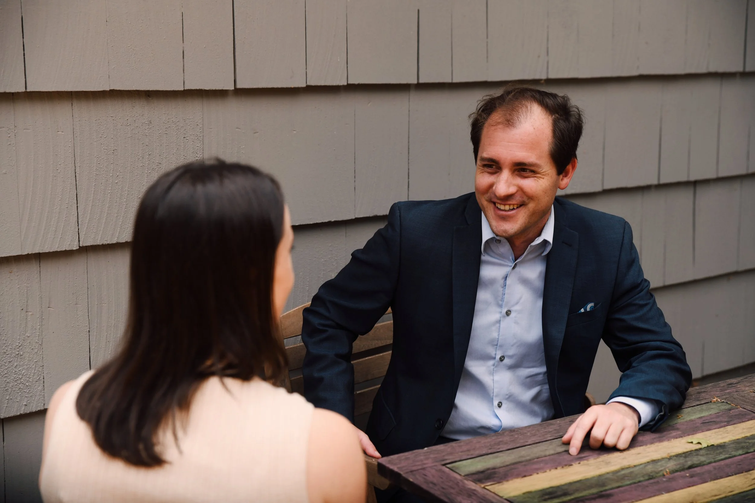 A man and a woman are sitting at a wooden picnic table outdoors, engaging in a conversation. The man is smiling and dressed in a navy blue suit and white shirt. The woman has dark brown hair and is wearing a sleeveless top. They are seated against a background of a gray wooden wall.