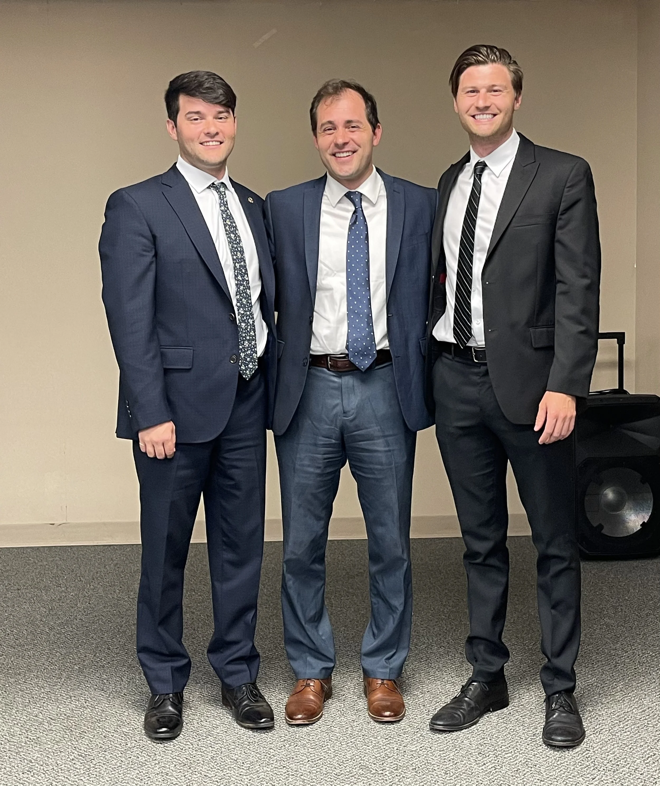Three men dressed in suits standing together indoors, smiling at the camera.