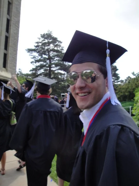 A young man in graduation cap and gown wearing reflective sunglasses, smiling at the camera, during a graduation ceremony with other graduates in caps and gowns outside.