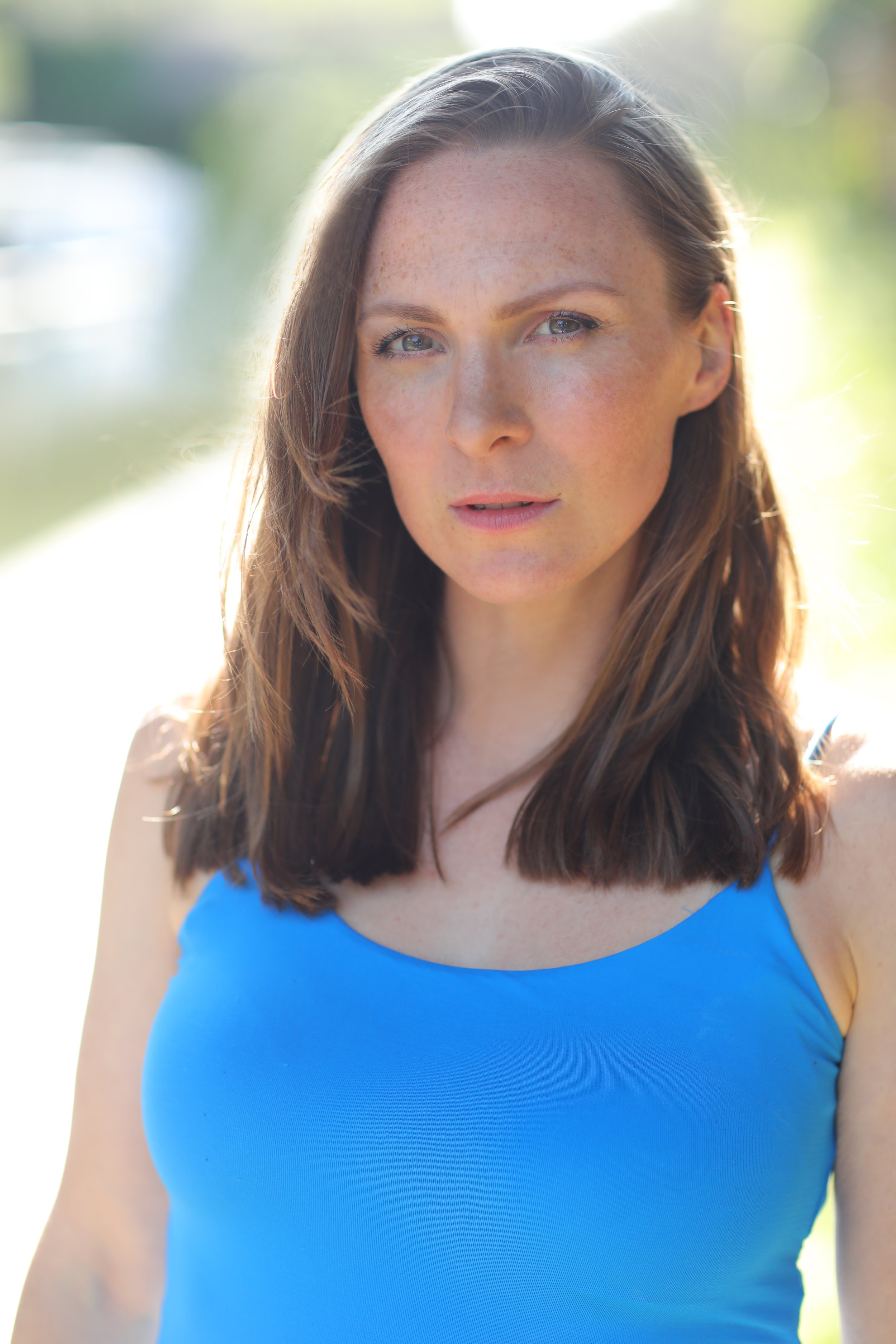 Close-up of a young woman with long brown hair, blue eyes, light makeup, and freckles, wearing a bright blue sleeveless top outdoors with a blurred green background.