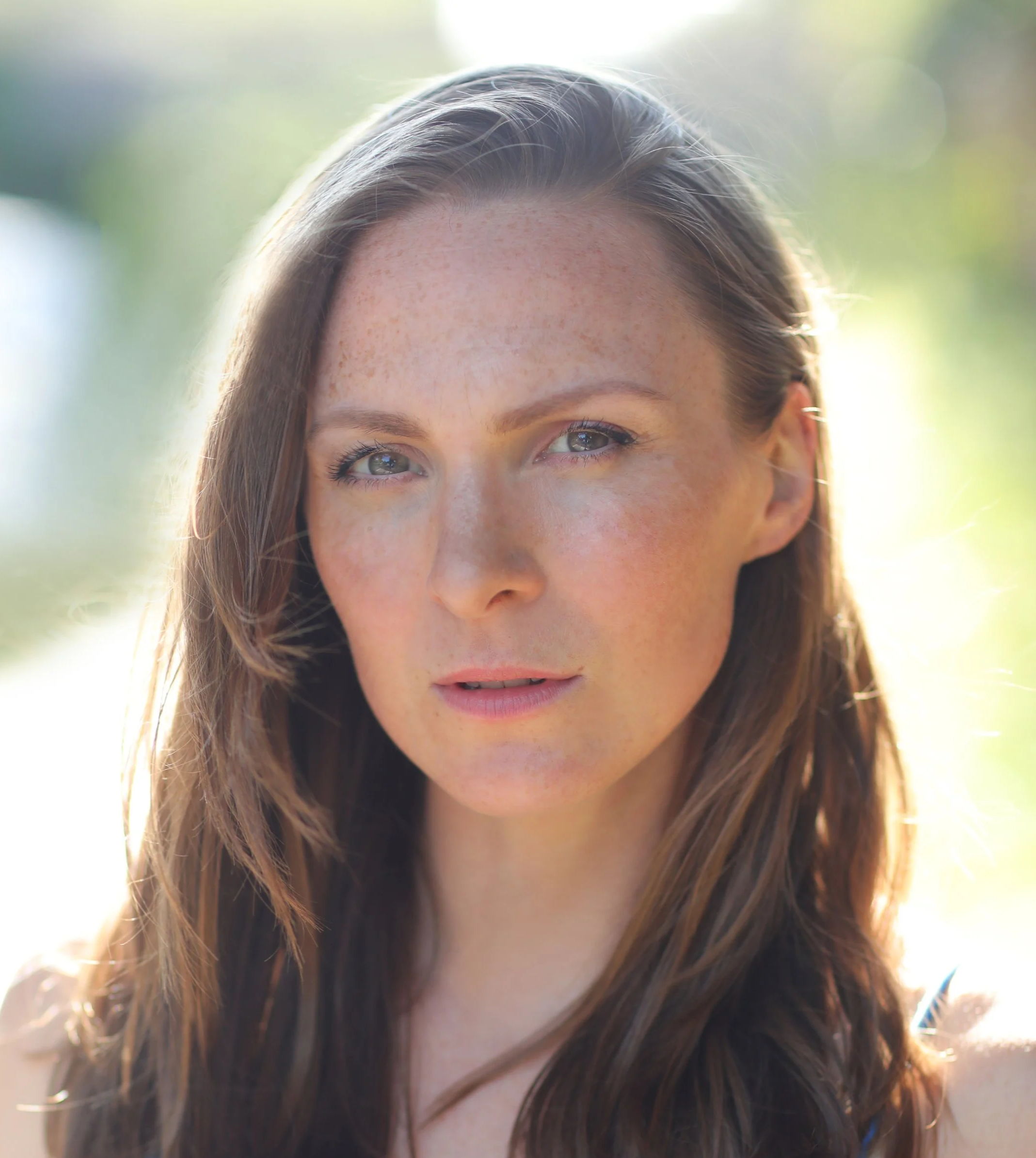 Close-up of a woman with freckles and brown hair, smiling outdoors with sunlight highlighting her hair.