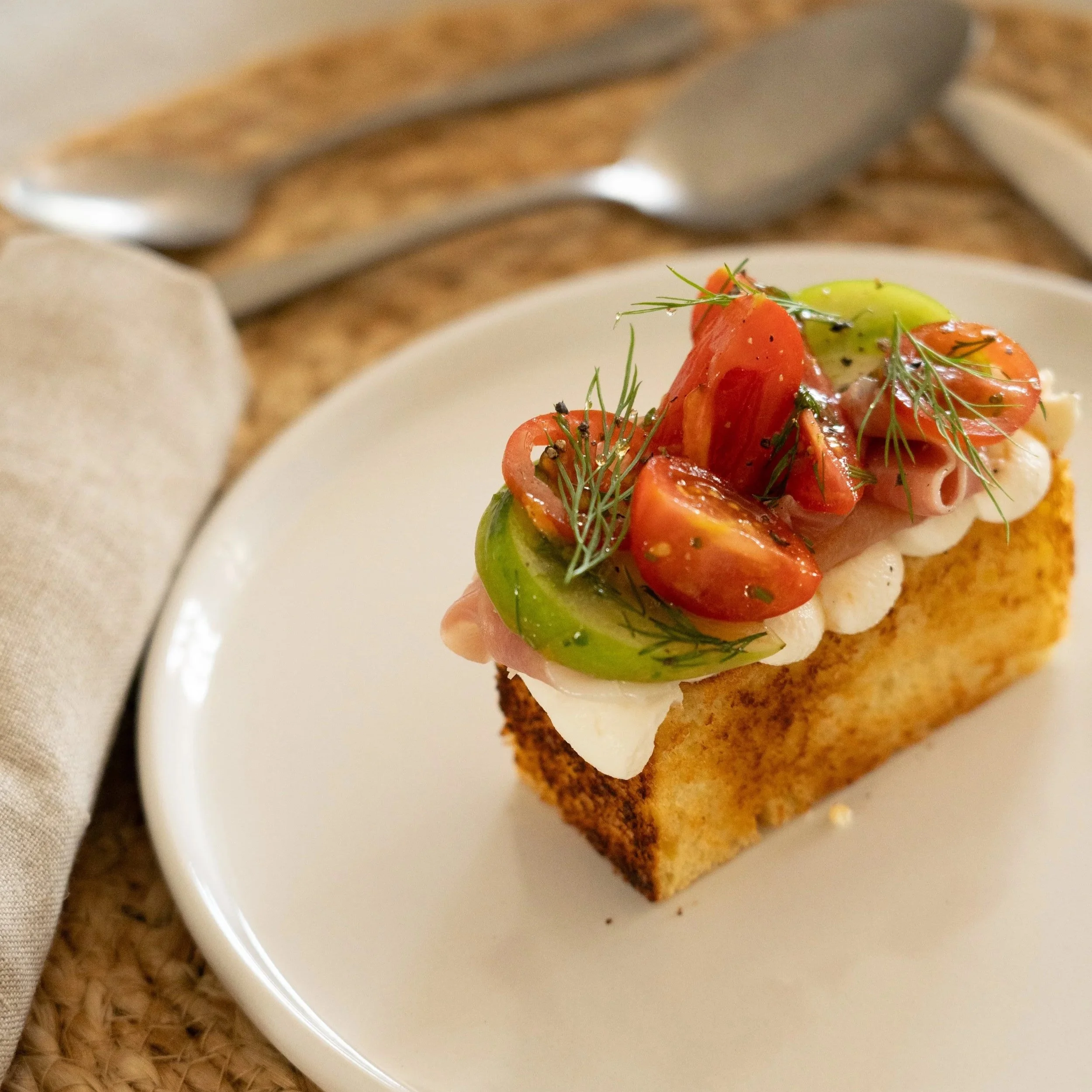 A piece of toasted bread topped with slices of tomato, avocado, and garnished with fresh herbs, served on a white plate.
