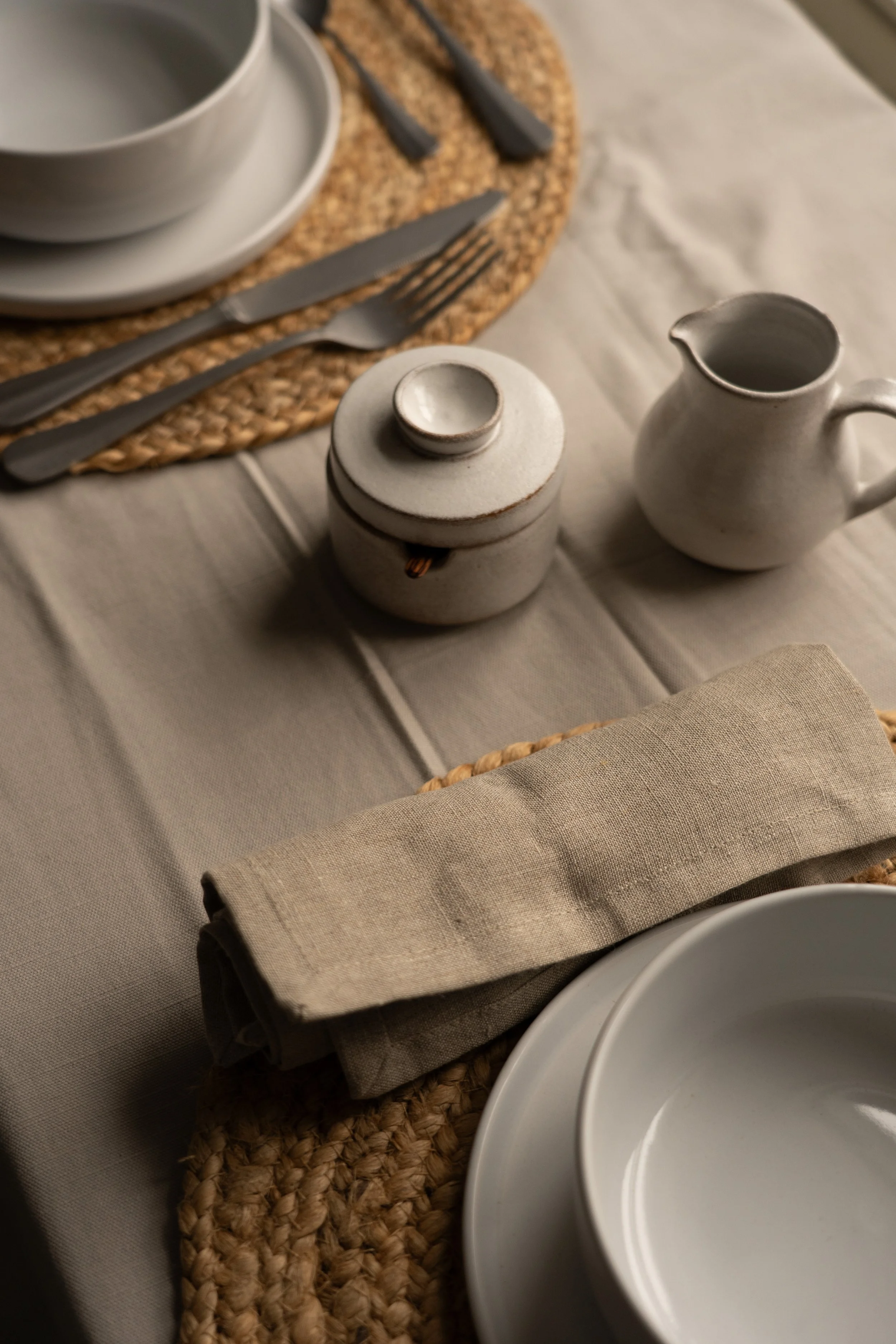 A table setting with a woven placemat, white ceramic bowls and plates, a small white ceramic pitcher, a linen cloth and napkin, and silverware including a knife and fork, set up for a private brunch in Devon