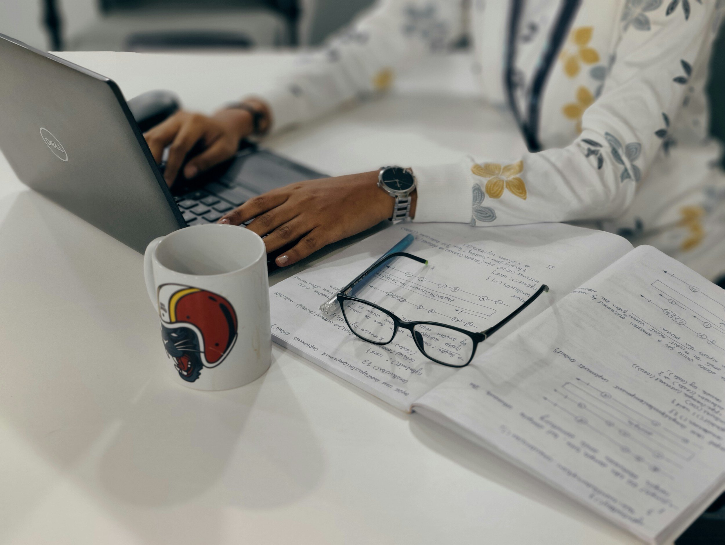 A person's hands typing on a Dell laptop keyboard on a white table. The person wears a floral-patterned shirt and a wristwatch. There are handwritten notes, a notebook, a pair of glasses, and a white mug with a logo on it on the table.