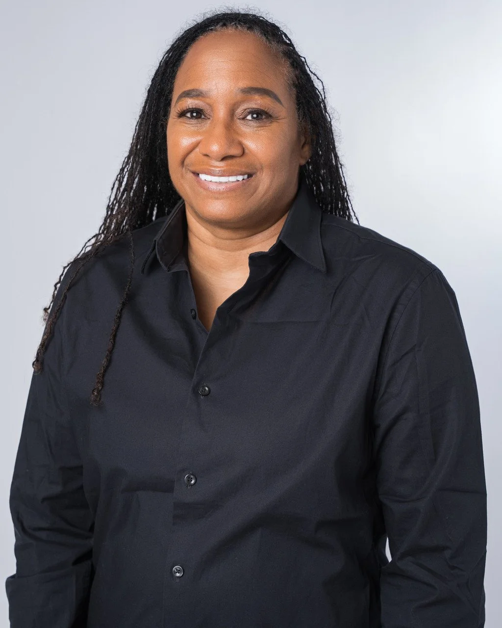 Portrait of a smiling woman with dark skin and long dreadlocks wearing a black button-up shirt against a plain gray background.