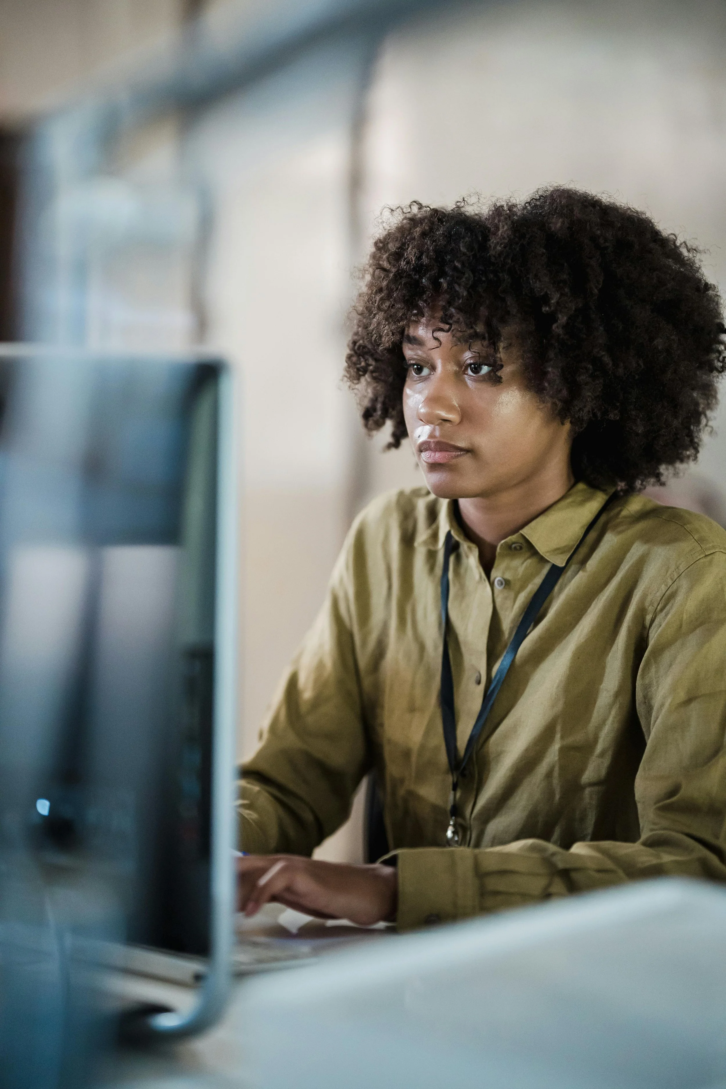 A woman with curly hair wearing a khaki shirt working at a desk, seen through a blurred computer monitor in the foreground.
