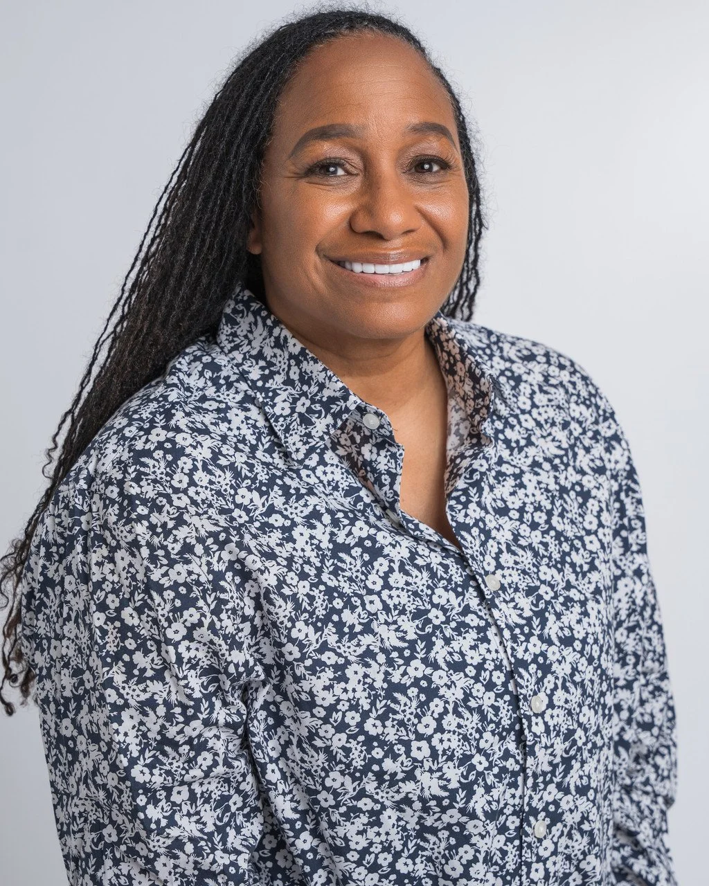 A woman with long dreadlocks wearing a navy blue and white floral blouse, smiling and looking at the camera against a plain light gray background.