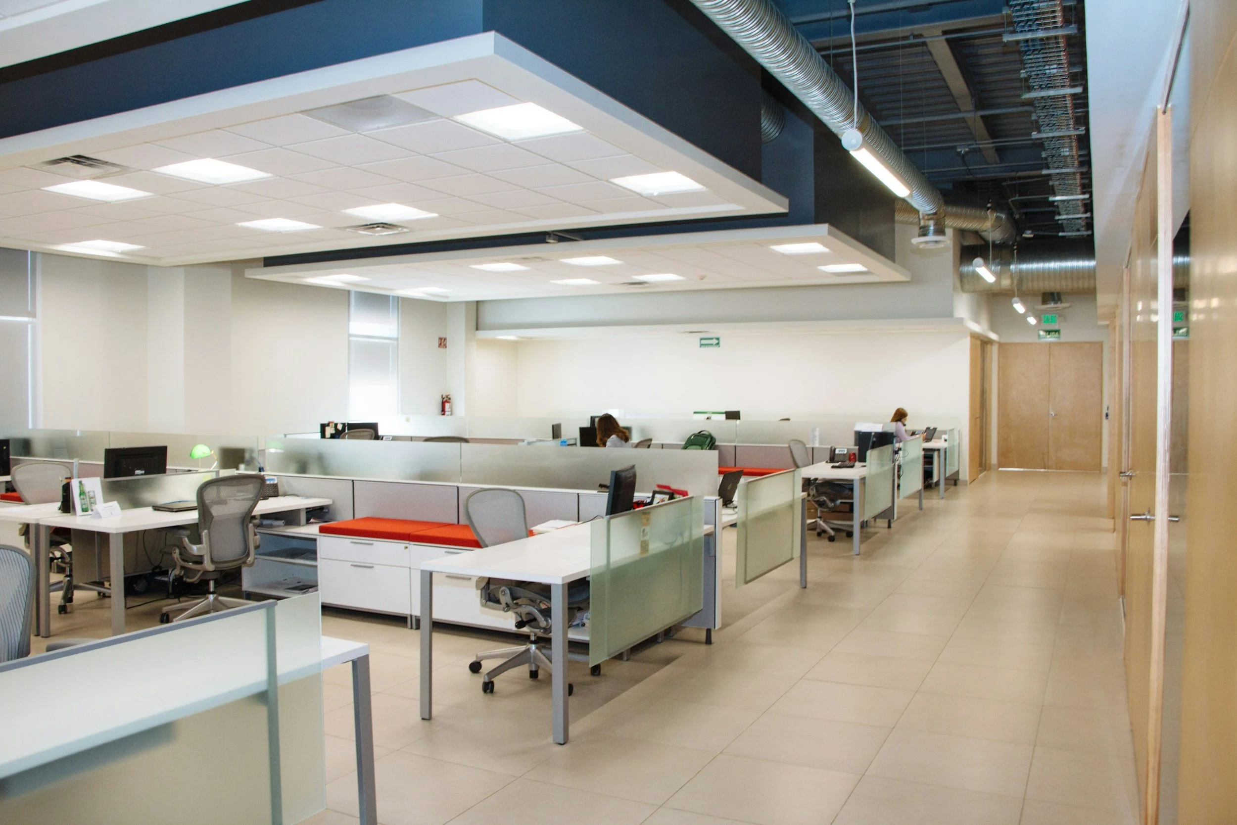 Modern open-plan office with cubicles, desks, chairs, computers, and large windows. Exposed ceiling ductwork and lighting fixtures are visible.