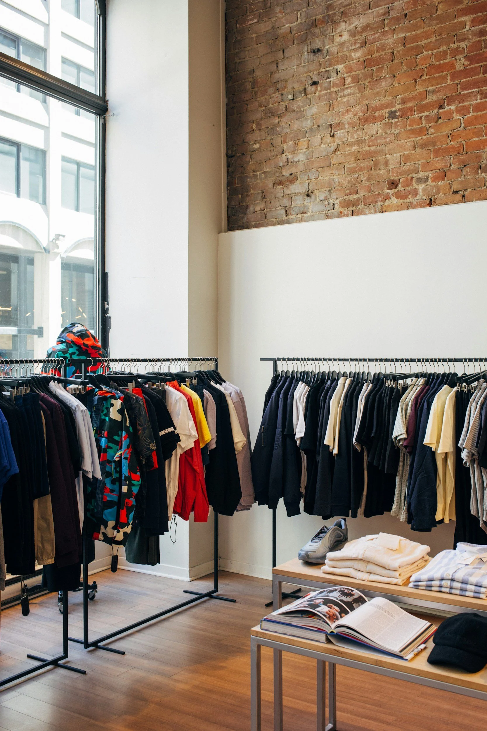 Clothing store with racks of casual and athletic wear, a pair of sneakers, and folded shirts on tables, with exposed brick wall and large windows letting in natural light.