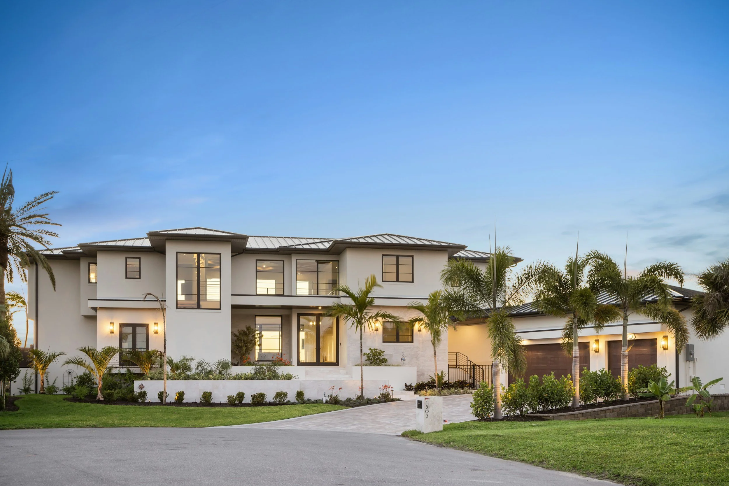 Modern white house with large windows, surrounded by palm trees and a well-maintained lawn, under a clear blue sky at sunset.