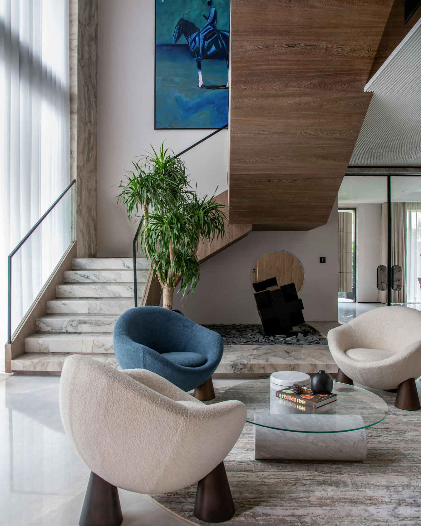 Modern living room with marble stairs, large green plant, blue armchair, white and beige chairs around a marble coffee table, colorful abstract painting of a horse on the wall, and large windows with curtains.
