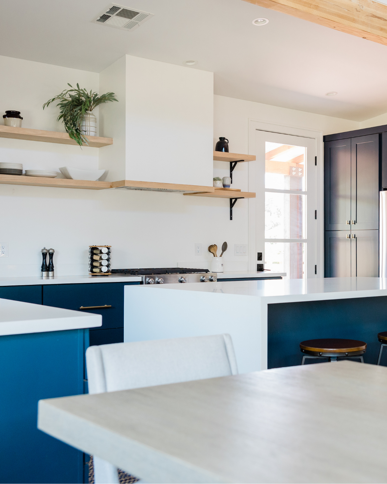A modern kitchen with blue lower cabinets, white countertops, and open wooden shelves on white walls. There is a stove, a white container with utensils, and a door leading outside. A black cabinet or pantry is visible on the right.