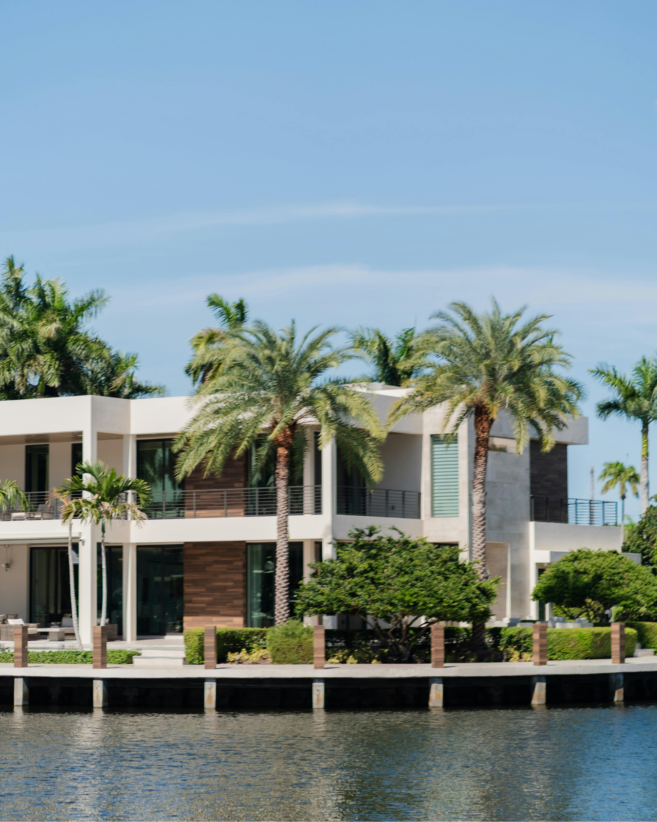 Modern house with large windows and multiple balconies, surrounded by palm trees, situated along a waterway under a clear blue sky.