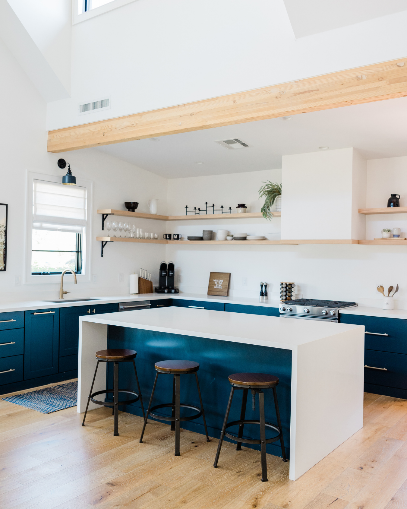 Modern kitchen with blue lower cabinets, white countertop, island with seating, open shelving with dishes, and wooden accents.