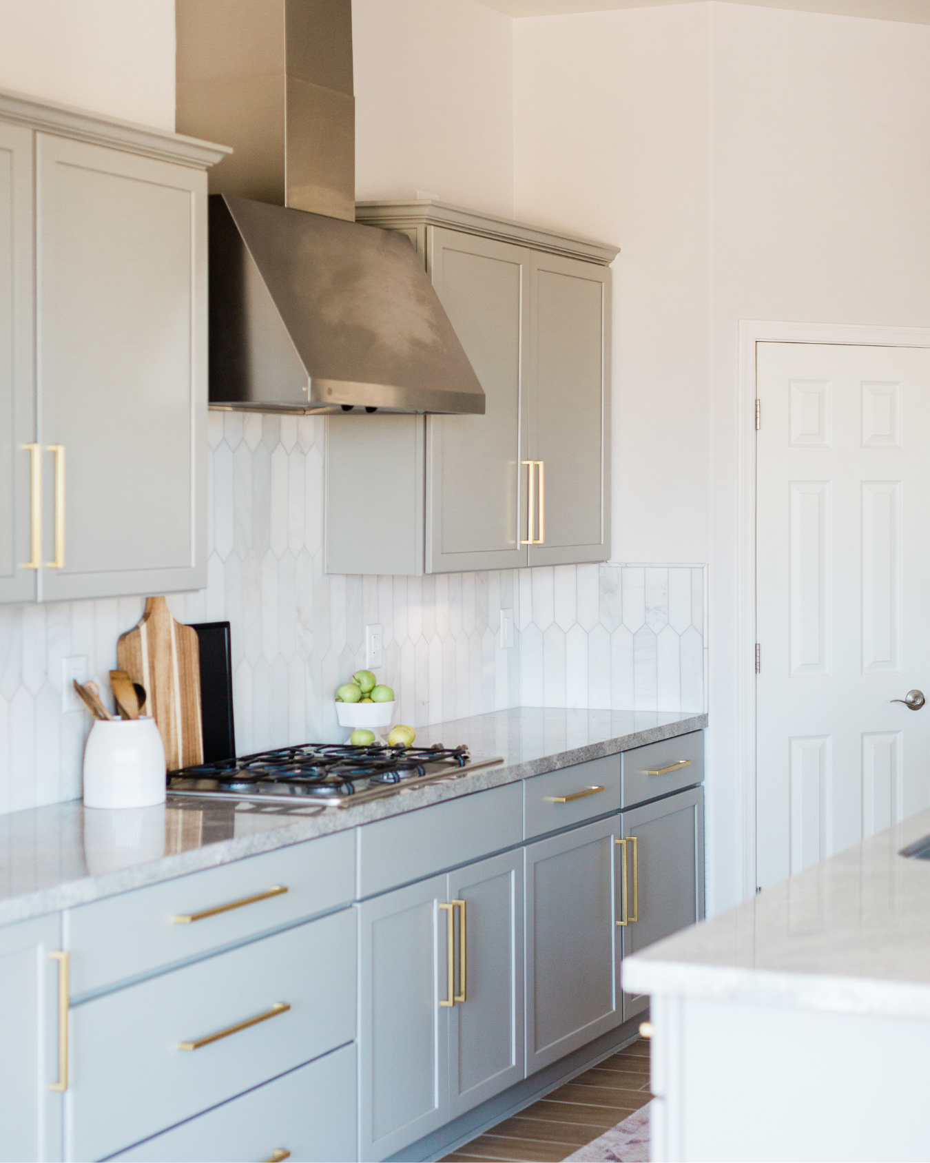 A modern kitchen with light gray cabinets with gold handles, a countertop with a bowl of green apples, a cutting board, and a white container holding wooden utensils, a gas stove with black grates, a stainless steel range hood, a white tiled backsplash with a hexagon pattern, and a closed white door.