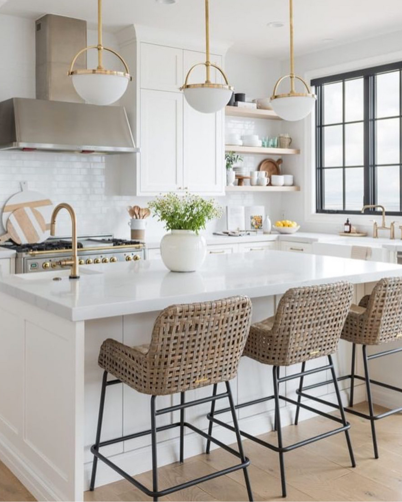 Bright, white kitchen with a large island, beige woven bar stools, open shelving with dishes, a window with black framing, and pendant lights hanging over the island.