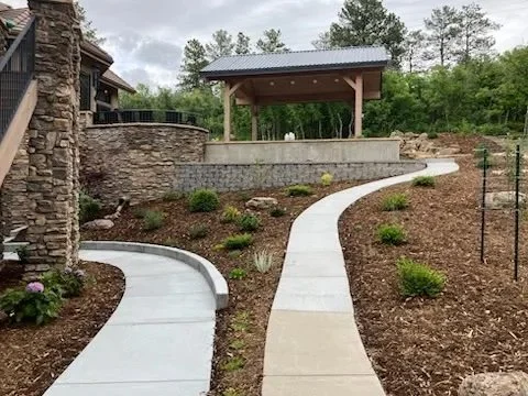 Curved concrete walkway in a landscaped yard leading to a stone and wood pavilion with a metal roof. The yard has mulch and small green plants.