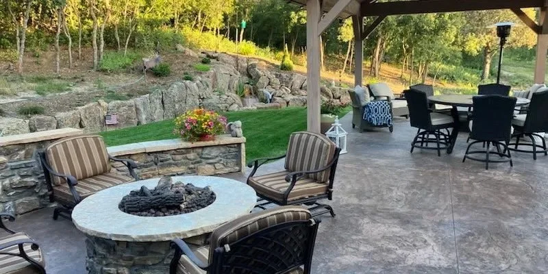 Outdoor patio with stone fire pit, patio table and chairs, covered seating area, and a landscaped yard with trees and rocks.