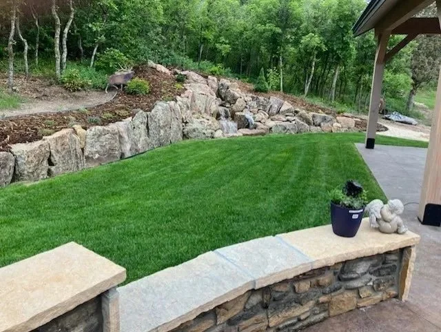 A backyard with a lush green lawn, stone border wall, and rocks in a wooded area. A deer is seen in the background near the trees.