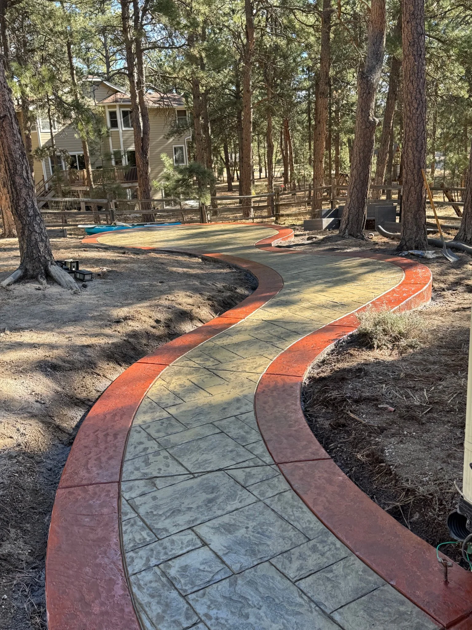 A winding concrete pathway with red border in a wooded backyard surrounded by tall pine trees, with a house in the background and some outdoor furniture and tools.