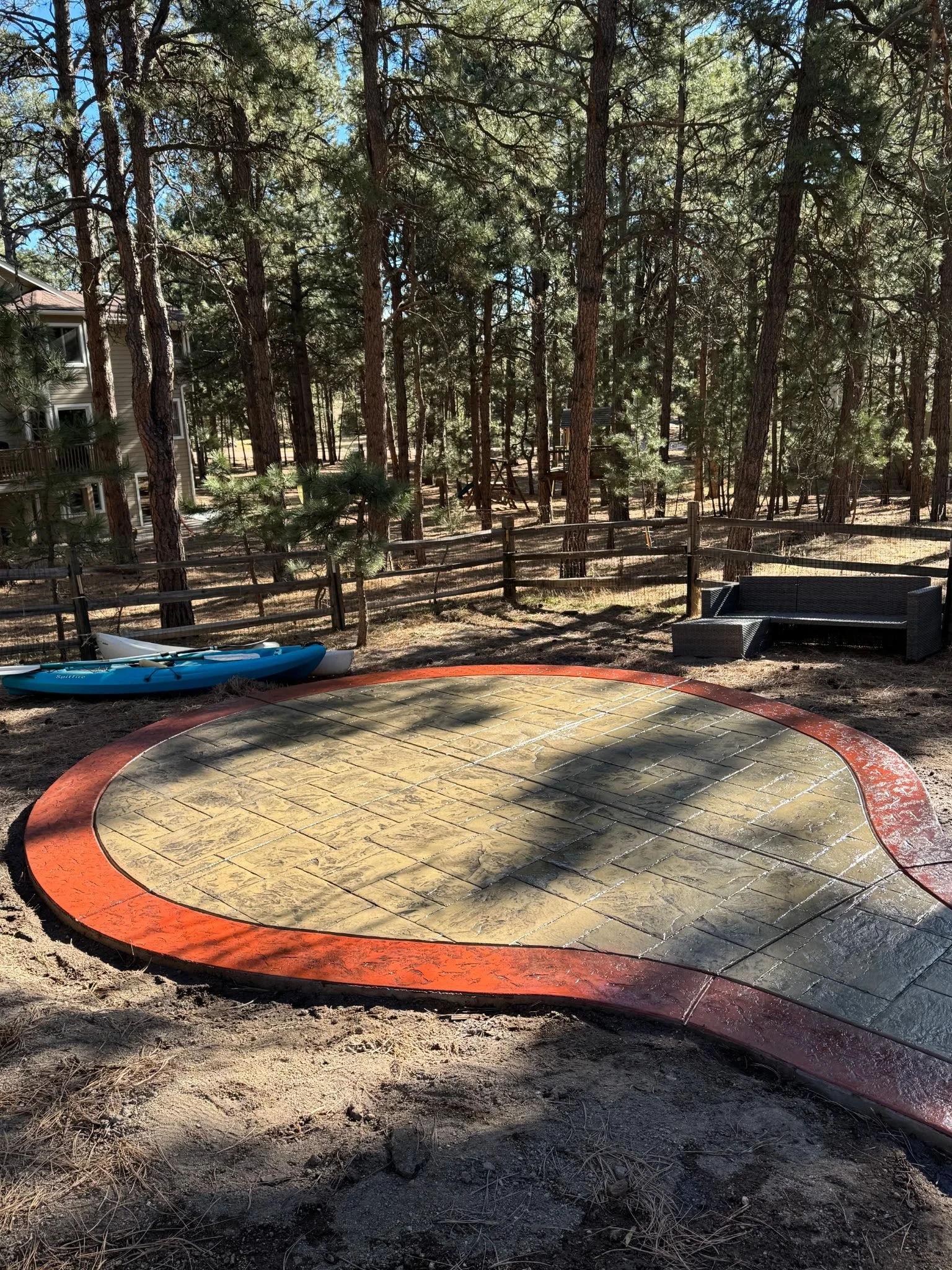 A backyard with a trampoline, a kayak, a black outdoor sofa, surrounded by tall pine trees and a wooden fence, with a house visible on the left side.