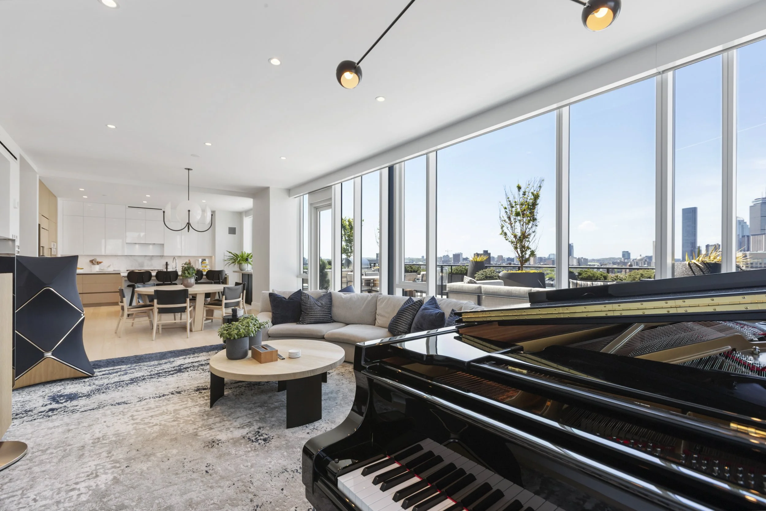 Modern living room with large floor-to-ceiling windows, a black grand piano, a white sectional sofa with pillows, a round coffee table, a patterned rug, and a dining area with chairs and a round table. There are plants and city views outside.