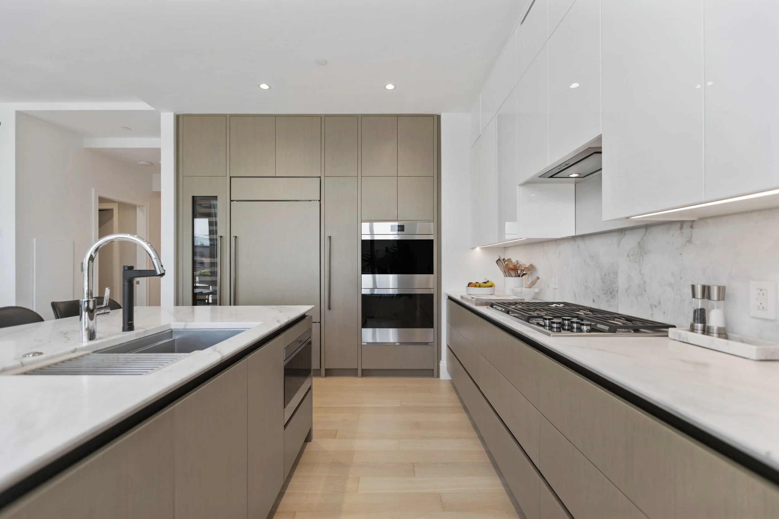 Modern kitchen with white marble countertops and light wood cabinetry, built-in oven, gas stove, stainless steel sink, and under-cabinet lighting.