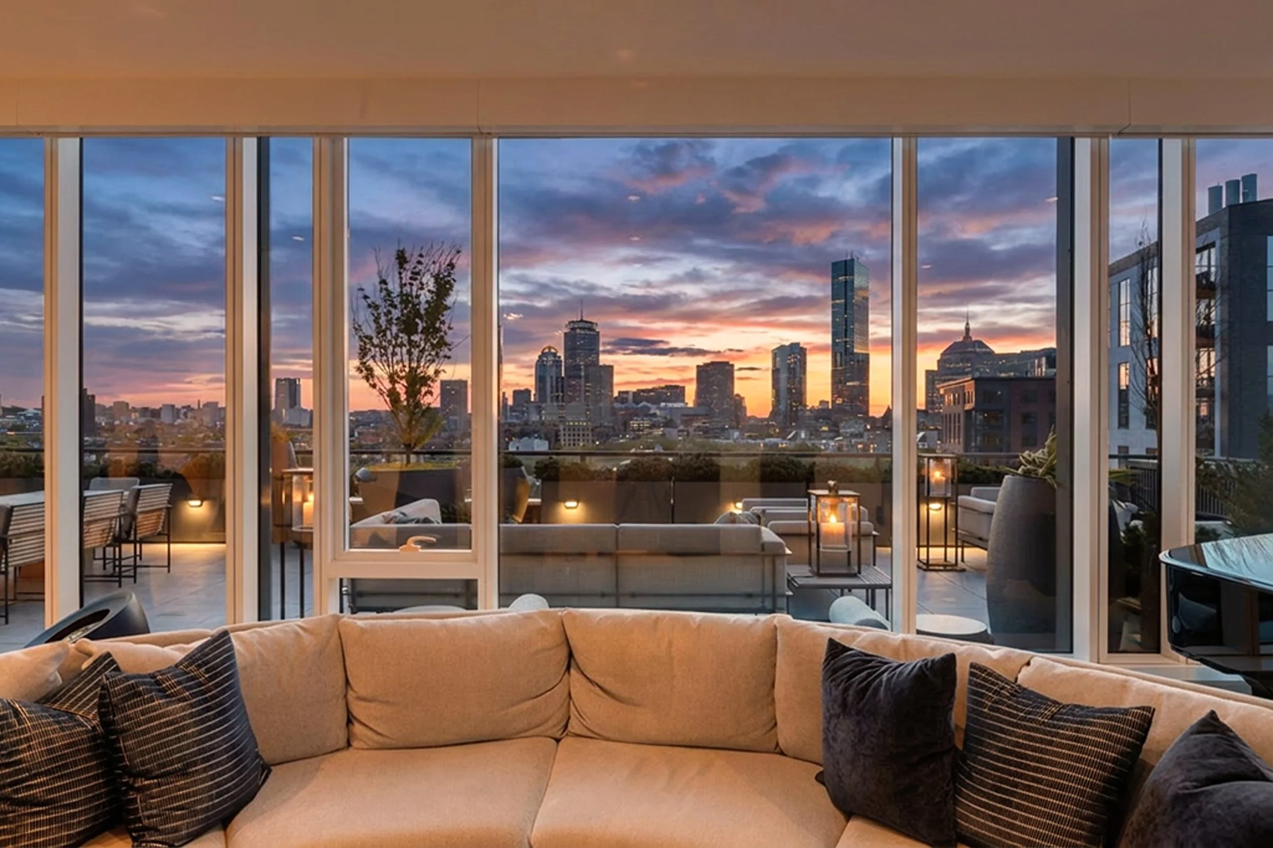 Living room with large glass windows showing a city skyline at sunset, furnished with a beige sofa and dark striped pillows.