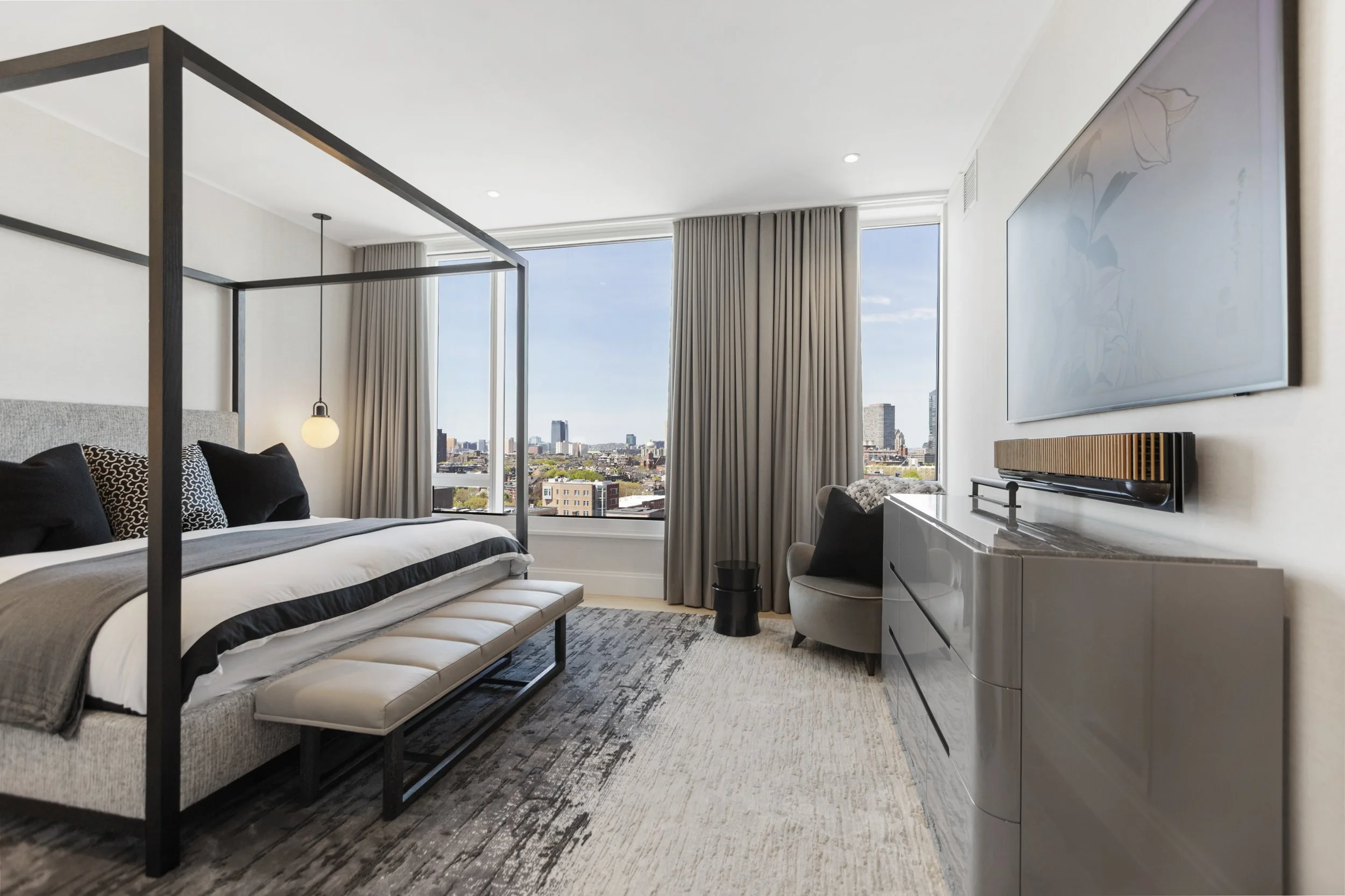 Modern hotel room with a large bed, a bench at the foot, a wall-mounted TV, a small seating area, and city skyline views through floor-to-ceiling windows.