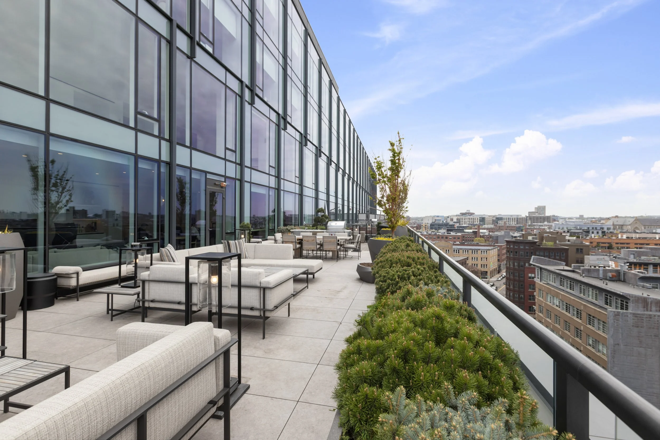 A modern rooftop terrace with outdoor seating, potted plants, and city views under a blue sky with clouds.