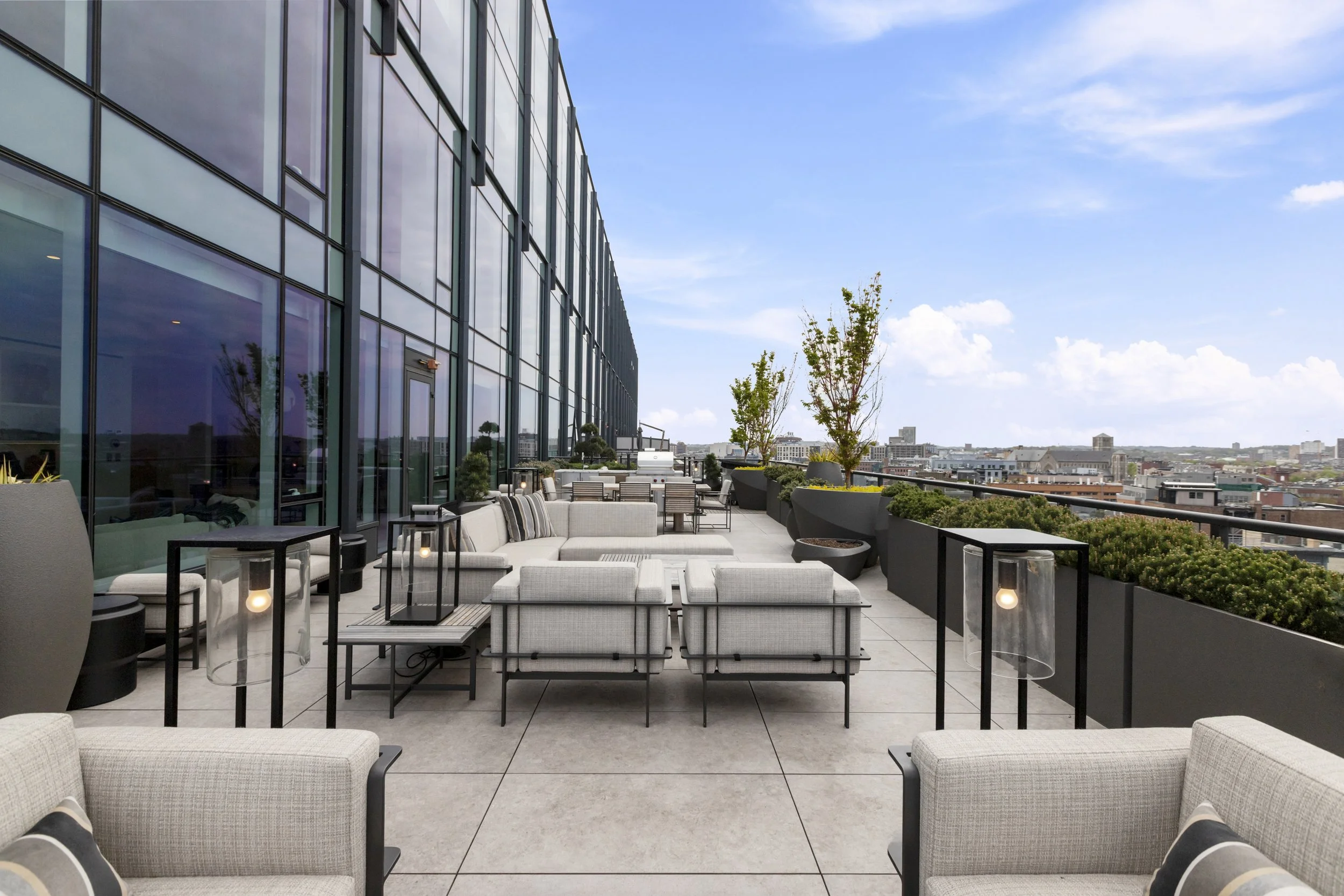 Rooftop terrace with modern outdoor furniture, potted trees, and cityscape view under a partly cloudy sky.