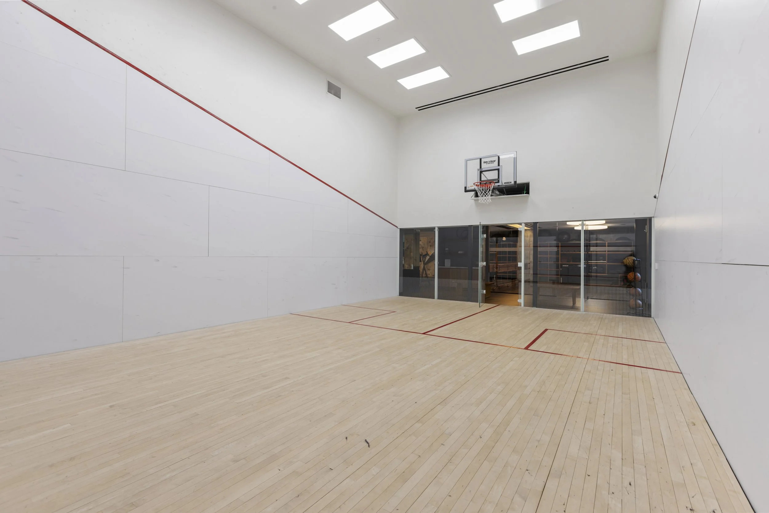 Indoor basketball court with light wood flooring, white walls, and a basketball hoop mounted on the wall. Large windows and ceiling lights provide lighting.