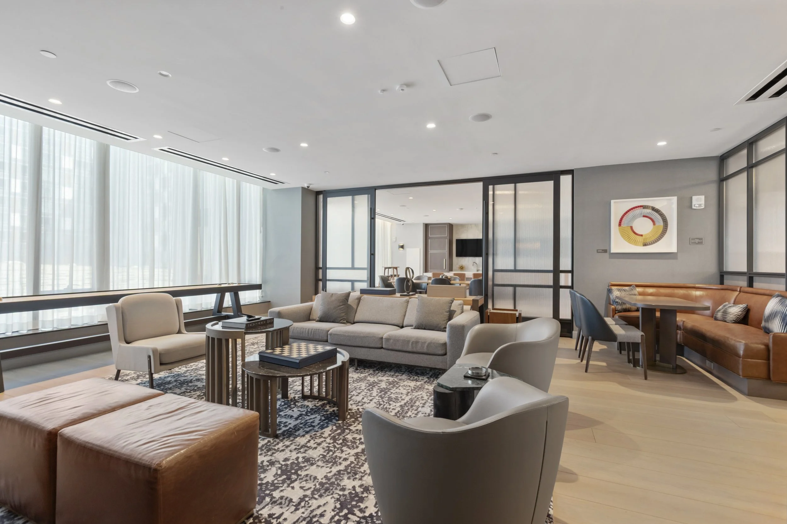 Modern hotel lobby with large windows, white curtains, and various seating including beige and brown chairs and sofas. Decor includes a patterned rug, a wall art piece, and a flat-screen TV in the background.