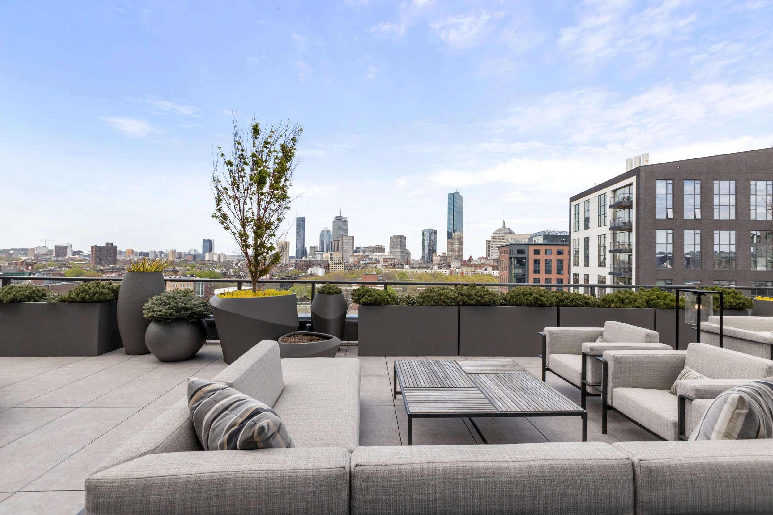 A modern apartment balcony with outdoor seating, potted plants, and a city skyline view featuring tall buildings and a blue sky with some clouds.