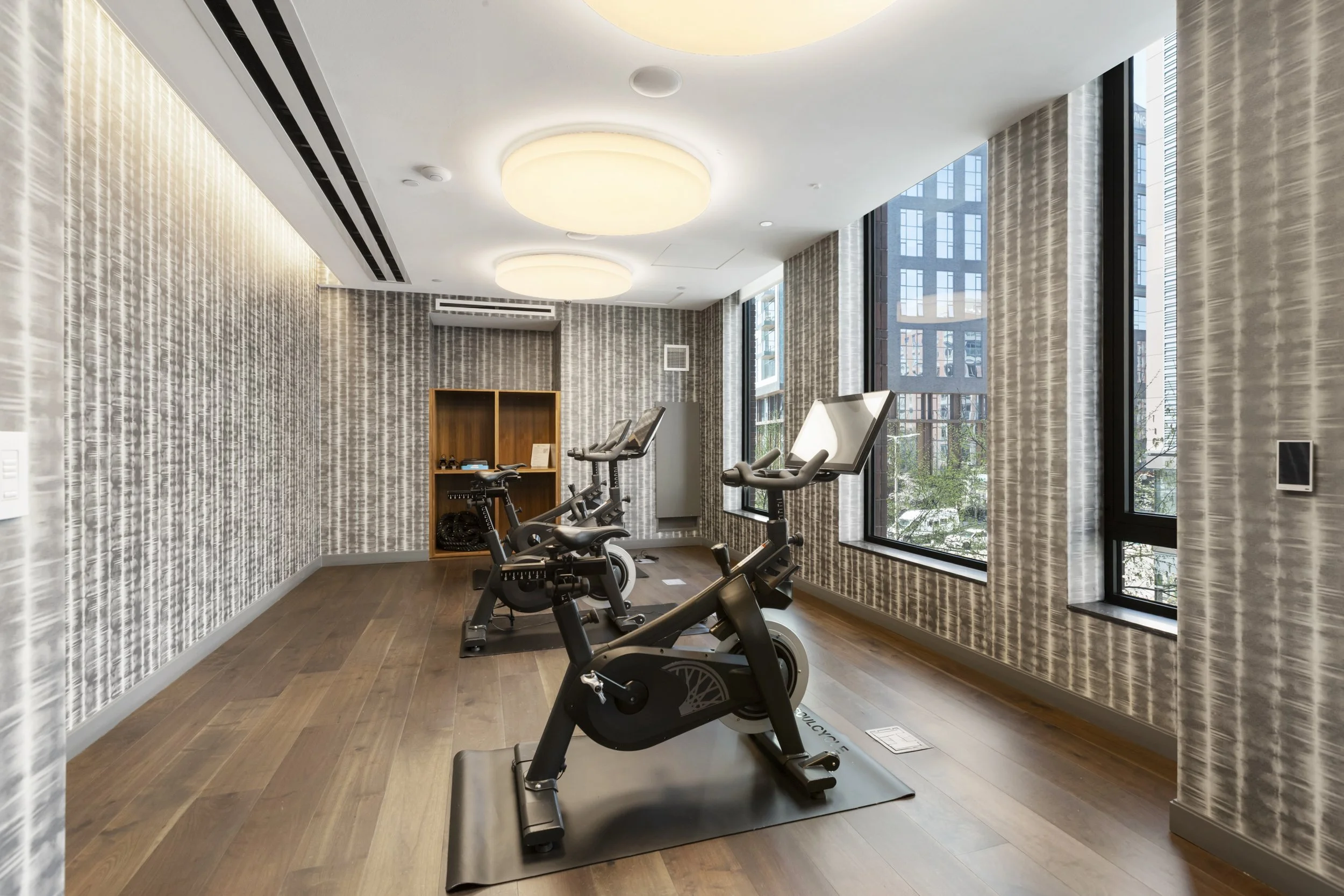 Indoor gym with three exercise bikes facing large windows, textured wallpaper, wooden floor, and ceiling lights.